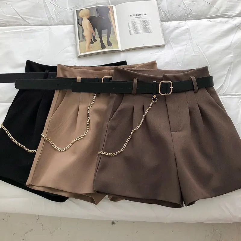 Two pairs of shorts, one black and one brown with a chain detail, on a white surface.