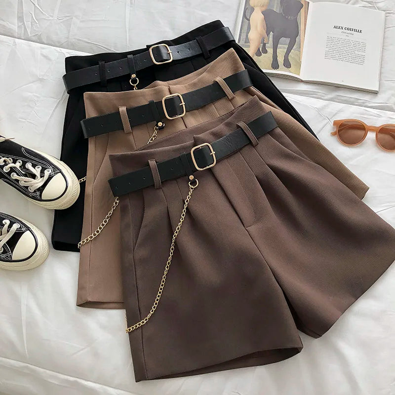 Three pairs of shorts with belts on a white surface with shoes and sunglasses.