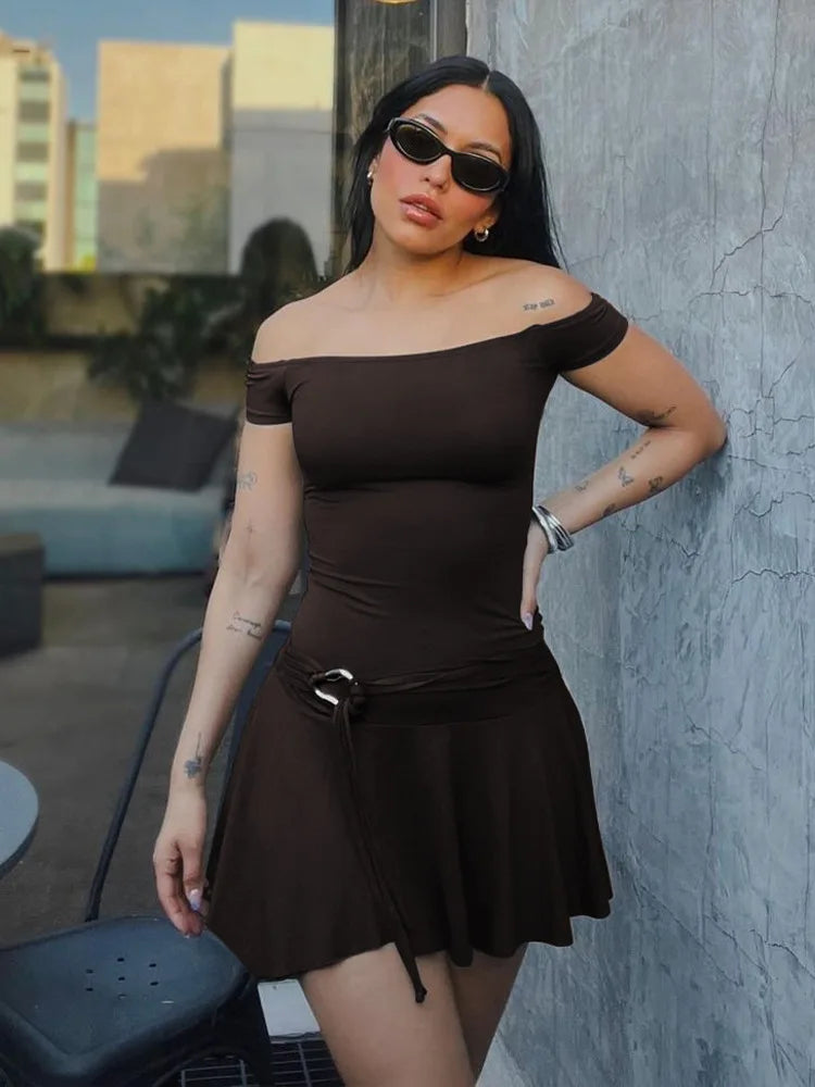 Woman in a brown off-shoulder dress standing against a textured wall.