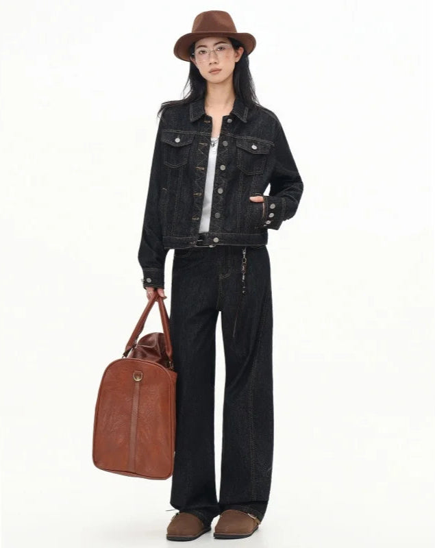 Person wearing a black denim jacket and pants, holding a brown leather bag on a white background