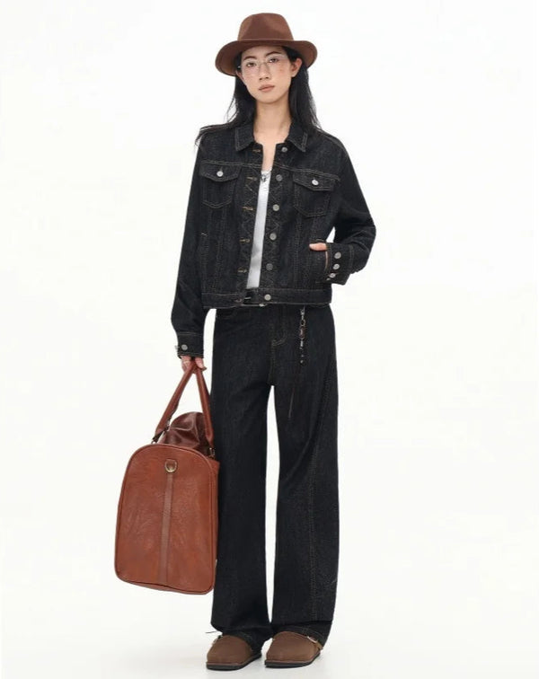 Person wearing a black denim jacket and pants, holding a brown leather bag on a white background