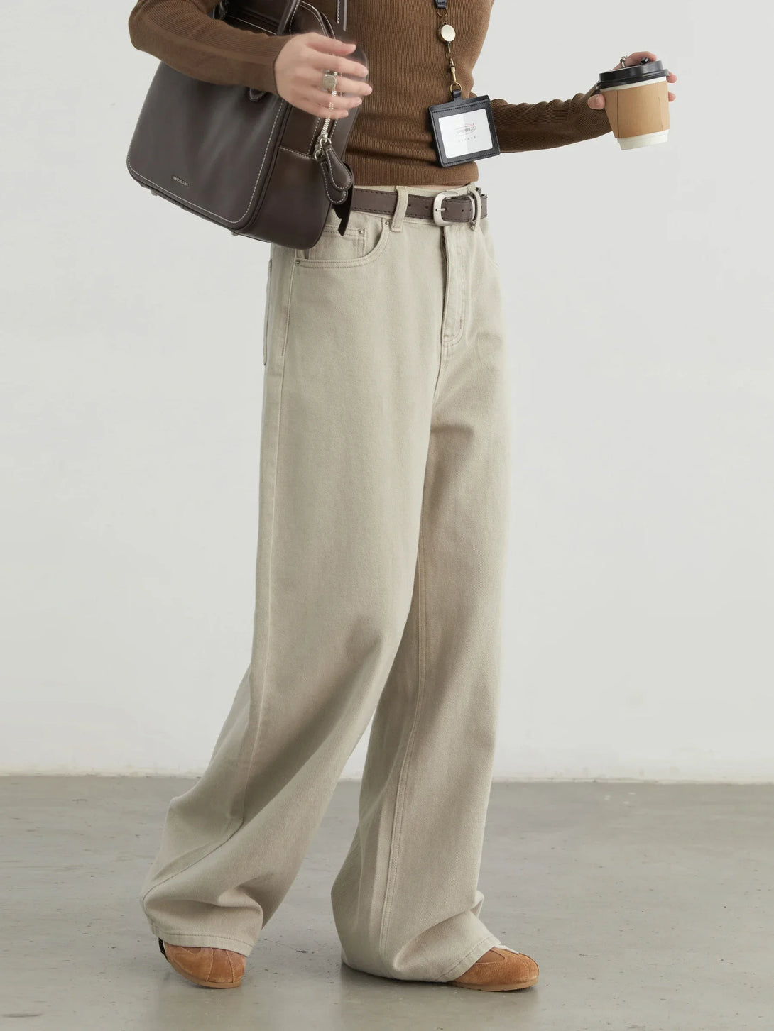 Person wearing a brown sweater, beige wide-leg pants, and holding a coffee cup against a white background